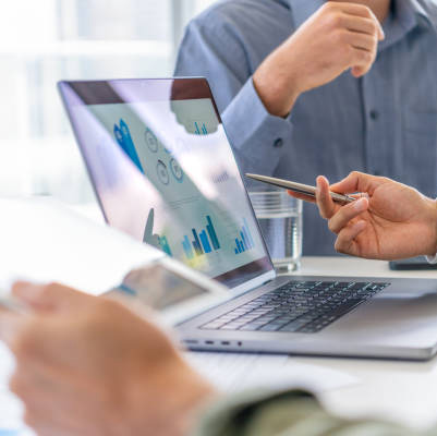 Group of men working with Paperwork on a board room table at a business meeting or seminar. The documents have financial or marketing figures, graphs and charts on them. They are all using laptop computers and digital tablets